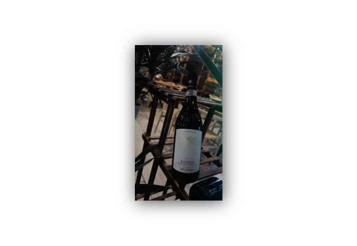 A bottle of 2014 Barolo wine labeled "Vinum Vita Est" and "Terre del Barolo" displayed on a wooden rack. The background features chairs and tables in an indoor dining setting, suggesting a restaurant atmosphere.