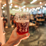 A hand holding a pint of amber beer in a glass with the Indie Beer Feast logo, positioned in the foreground. The background shows a softly lit festival hall with string lights, tables, and attendees, all slightly blurred to emphasise the beer in focus.