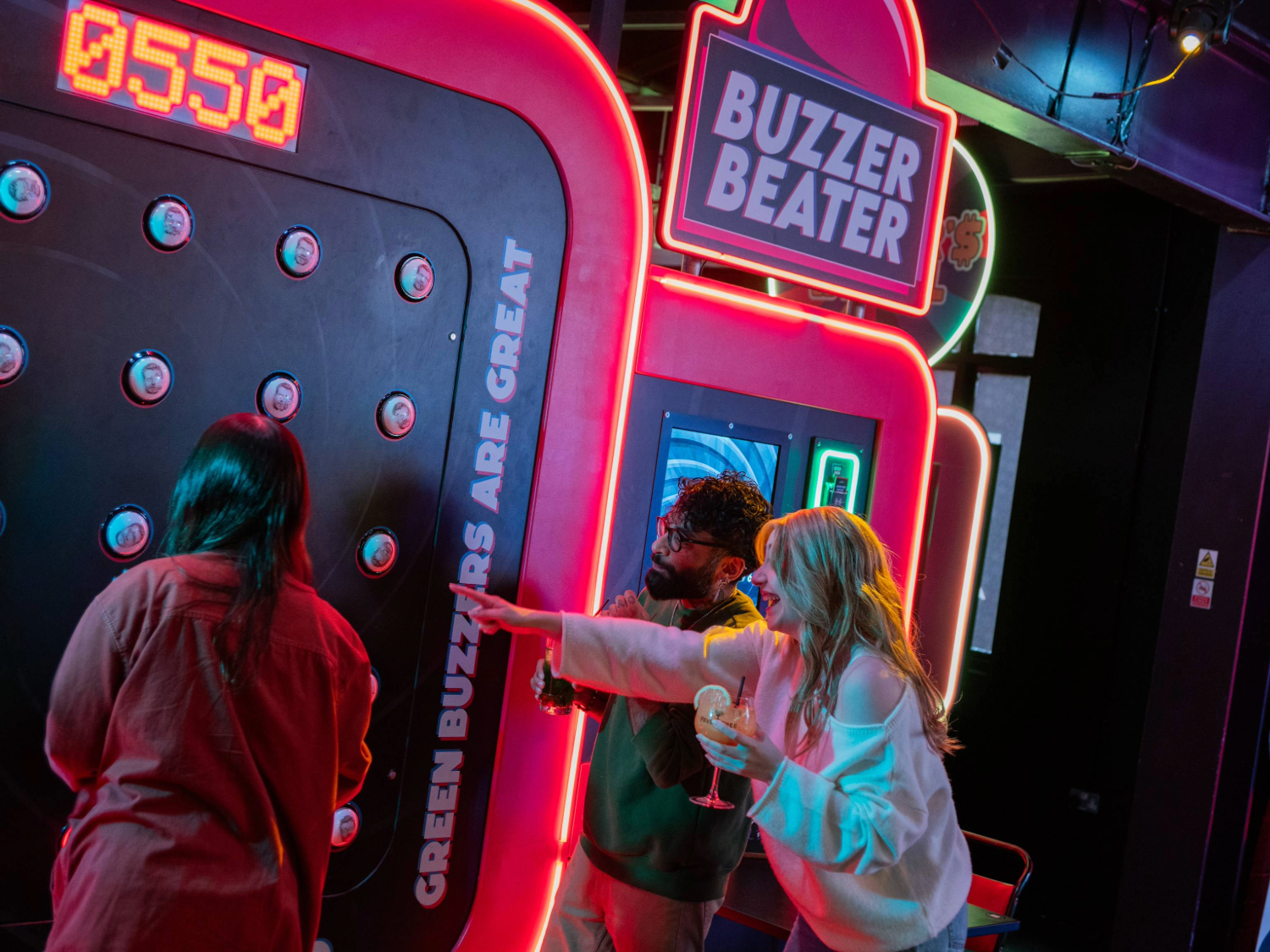 Three people playing an interactive arcade game called “Buzzer Beater” in a neon-lit venue. The game features a large vertical panel with multiple round buzzers and a digital score display reading “0550.” Bright signage on the machine includes phrases like “Green buzzers are great.” One person is actively playing, while another points at the panel, holding a drink. The setting is vibrant with colourful lights and a lively atmosphere.