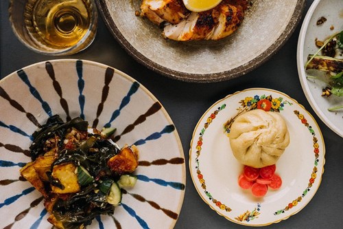 Top-down view of a table with assorted dishes, including a plate of roasted vegetables with greens, a decorative plate holding a steamed bun and pickled radishes, a stone bowl with grilled meat topped with a lemon wedge, and a glass of golden-coloured drink.