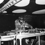 A black and white image of a DJ standing behind two record decks and a mixer, playing music.