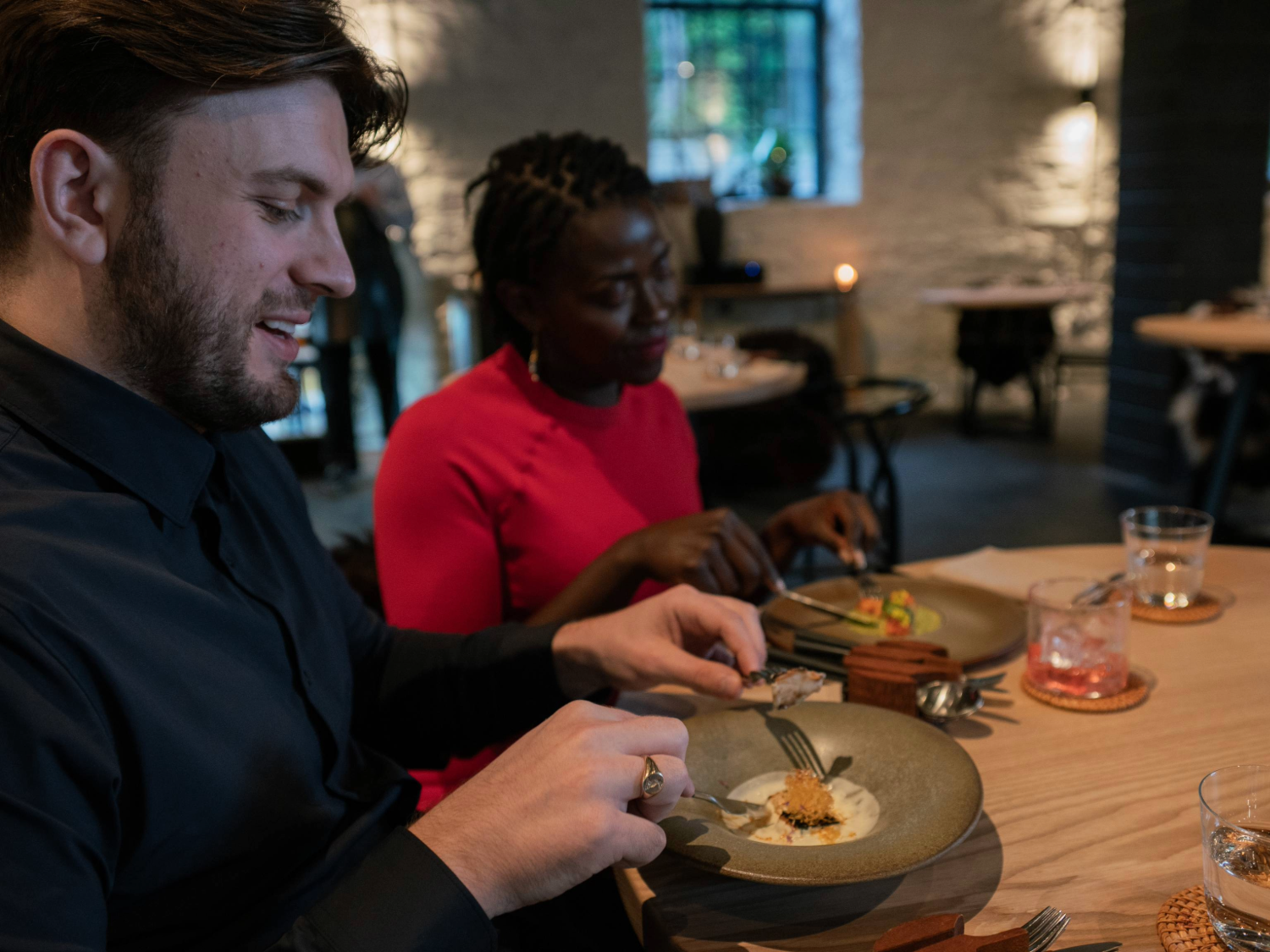 Two people seated at a wooden table in a modern restaurant, enjoying a plated dish. The table holds ceramic plates with artfully presented food, cutlery, and glasses with drinks. The background shows softly lit stone walls, other tables, and a large window, creating a warm and contemporary dining atmosphere.