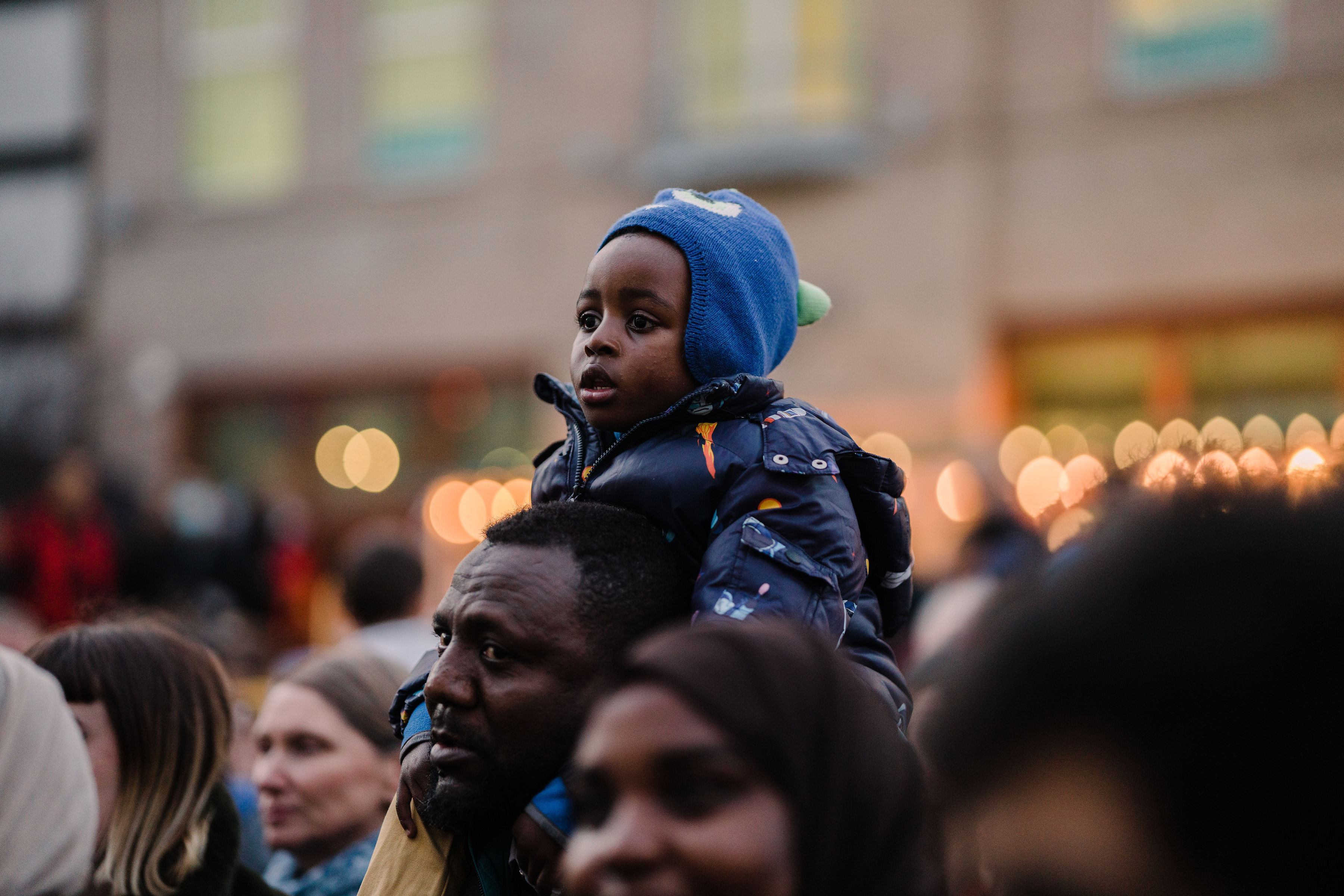 A child wearing a blue hat and padded jacket sits on someone’s shoulders in a crowded outdoor setting. The background shows blurred lights and a building, suggesting an event or gathering.