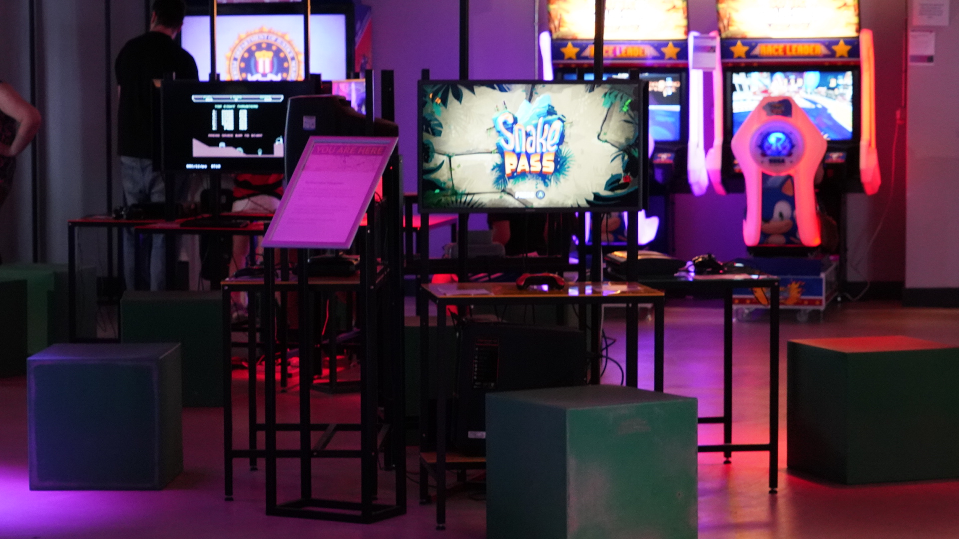 Gaming area with a monitor displaying the game ‘Snake Pass’ on a central table, surrounded by green cube seats. In the background, several arcade machines with bright screens and colourful designs are visible, creating a vibrant atmosphere.