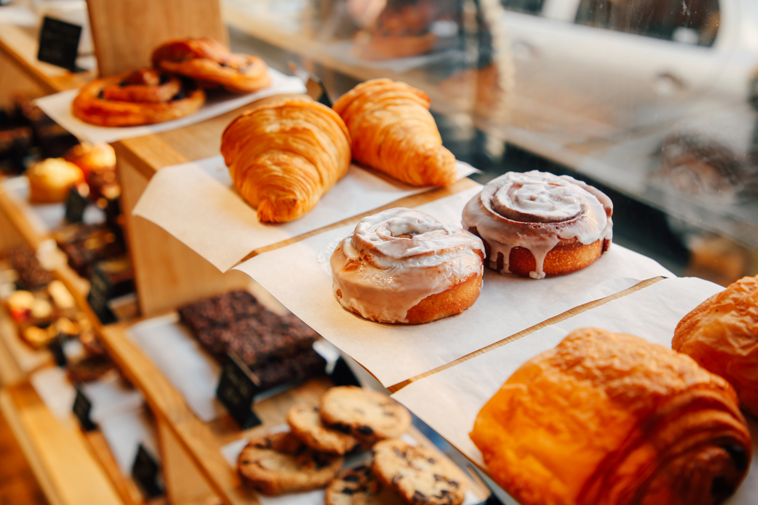 Bakery display with wooden shelves holding a variety of pastries. Visible items include golden croissants, iced cinnamon rolls, pain au chocolat, chocolate brownies, and cookies arranged on trays lined with parchment paper. Sunlight streams through the window, illuminating the baked goods.