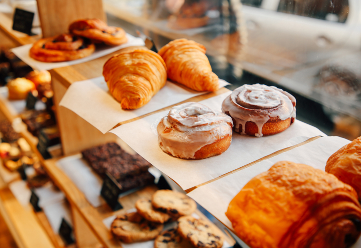 Bakery display with wooden shelves holding a variety of pastries. Visible items include golden croissants, iced cinnamon rolls, pain au chocolat, chocolate brownies, and cookies arranged on trays lined with parchment paper. Sunlight streams through the window, illuminating the baked goods.