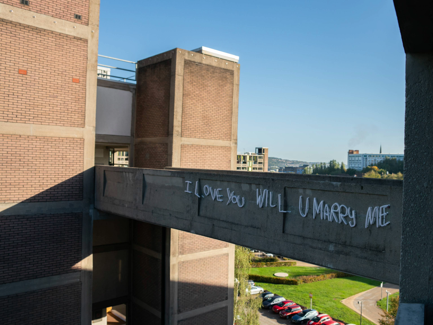A concrete bridge connects two section of an apartment block. The words 'I love you will u marry me' have been spray painted on the side of the bridge. They have since been picked out in neon lighting.