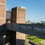 A concrete bridge connects two section of an apartment block. The words 'I love you will u marry me' have been spray painted on the side of the bridge. They have since been picked out in neon lighting.