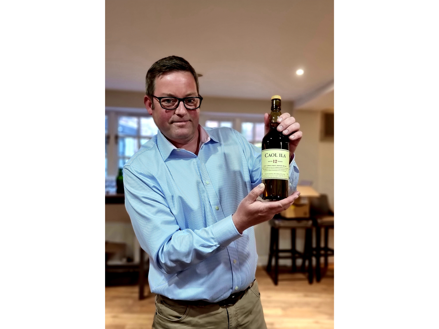 A person wearing a light blue, long-sleeved shirt is holding a bottle of Caol Ila 12-year-old single malt Scotch whisky with both hands. The bottle has a cream label with black and green text. The setting appears to be indoors, with wooden chairs and tables in the background and soft lighting from windows.