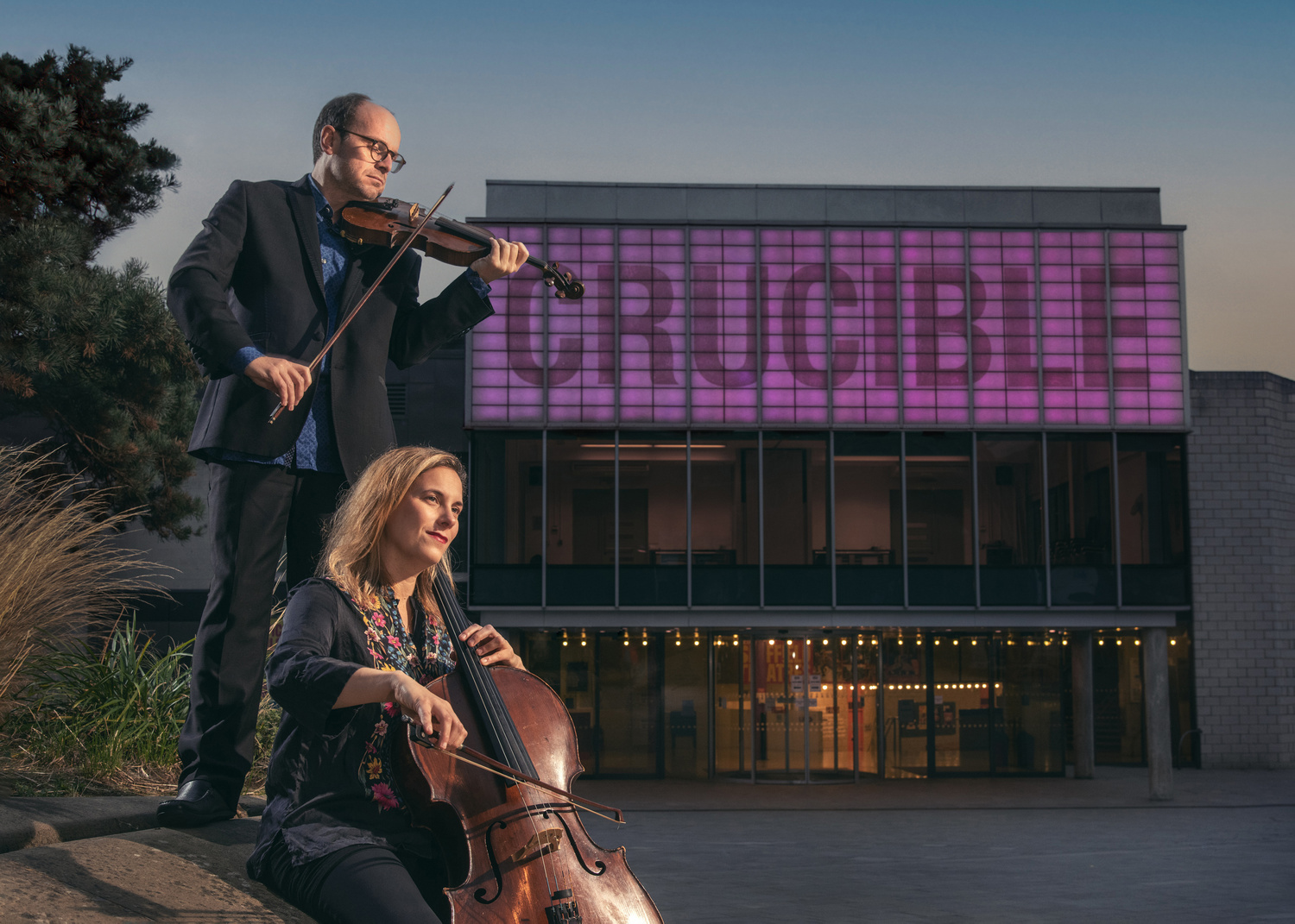 In Tudor Square, outside the Crucible Theatre, a woman plays a cello and a man plays a violin at dusk. 