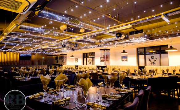 Elegant dining space with long rectangular tables set for a formal event. Each table is arranged with neatly folded cream napkins, polished glassware, and gold-colored cutlery. The ceiling features exposed ductwork and is adorned with strings of warm fairy lights, creating a festive atmosphere. Pendant lights hang above, and the walls display framed artwork. The overall setting is modern and sophisticated with a warm golden glow.