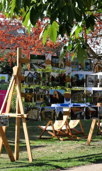 An outdoor art display at a garden event features rows of photographs or paintings mounted on wooden easels under a tree with autumnal leaves. Several deck chairs are arranged on the grass, and white marquee tents in the background host more visitors and exhibits. The scene is bright and sunny, with people browsing the artwork and enjoying the relaxed setting surrounded by greenery.