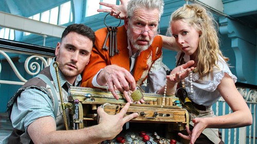 Three people wearing steampunk-style costumes are gathered around an elaborate wooden contraption decorated with brass fittings, keys, locks, and bottle caps. The setting includes ornate white railings and large windows in the background, suggesting an indoor venue with vintage architecture.