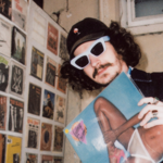 A man in sunglasses and a baseball cap holds a record sleevs. Behind him, the wall is covered in record sleeves on display.