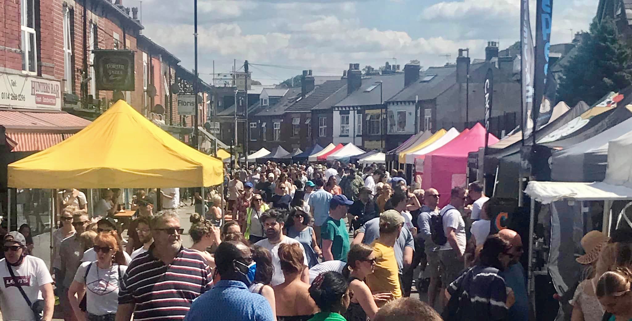 A busy summer street market with large crowds walking between stalls, including a bright yellow canopy, set along a row of shops on Sharrow Vale Road