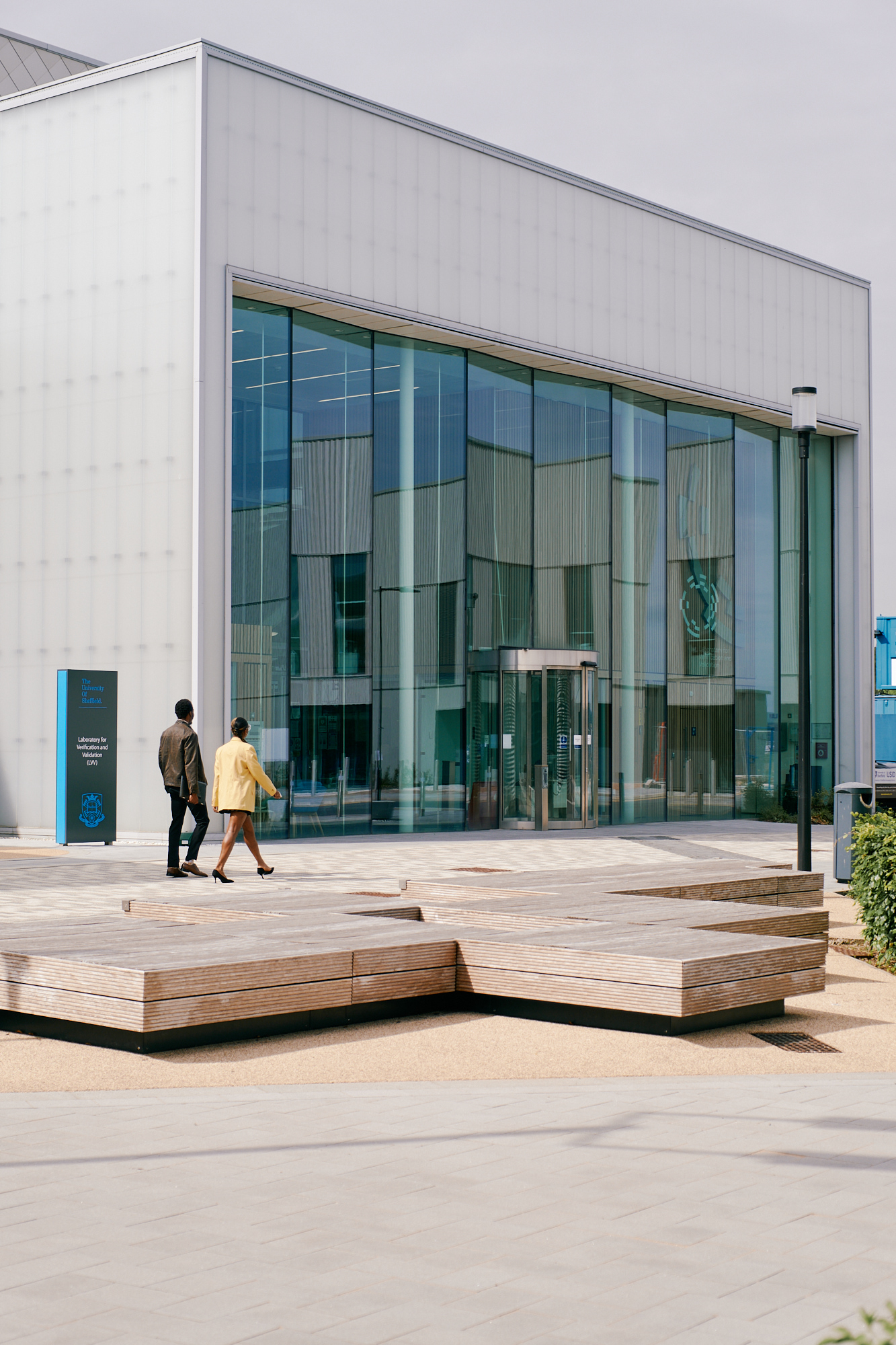 Exterior view of a modern building with large glass panels and a white, translucent facade. Two people walk toward the entrance, which features a glass door. In front of the building, there are geometric wooden benches on a paved area, with a blue signboard visible on the left side.
