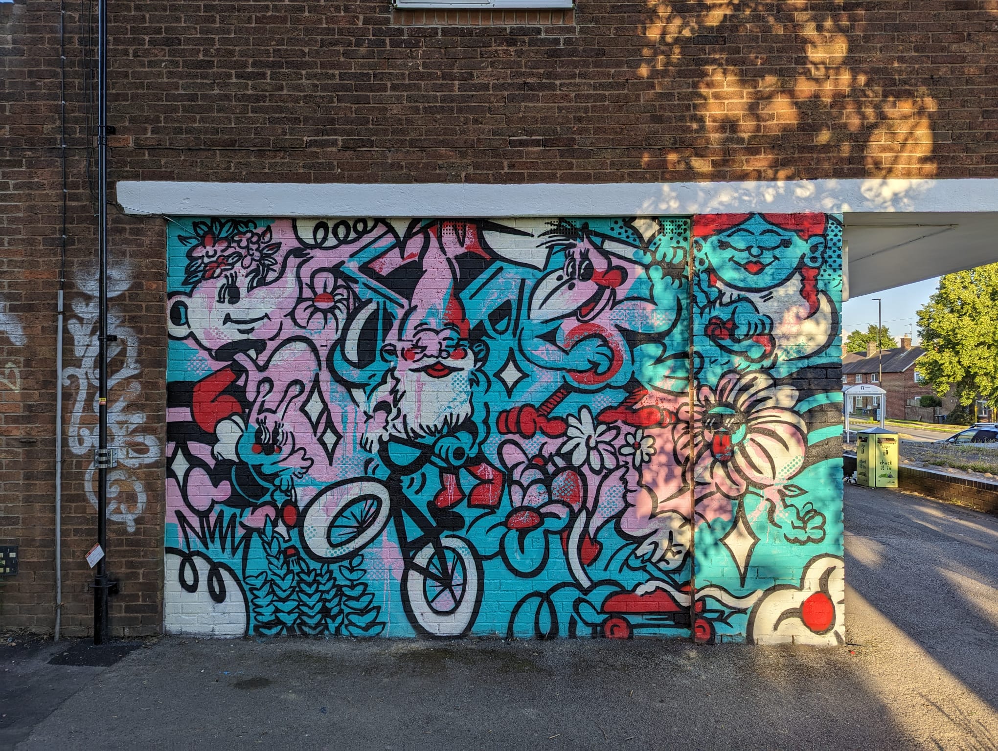 Colorful mural painted on a brick wall featuring a mix of cartoon-style characters, flowers, and abstract shapes in vibrant shades of red, turquoise, pink, and black. The design includes a central figure riding a bicycle, surrounded by playful patterns and decorative elements.
