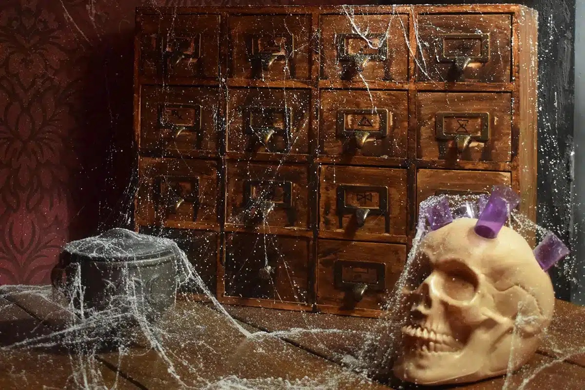 A dusty wooden cabinet with multiple small drawers is shown, covered in thick cobwebs. In front of it, on a worn wooden surface, lies a realistic skull prop with three purple crystal-like shards embedded in the top. To the left, there is a metal pot also draped in cobwebs. The background features a dark red patterned wall, creating an eerie, abandoned atmosphere suggestive of a spooky or mystical setting.