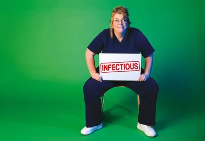 Nurse Georgie Carroll sits on a chair against a green background. She is wearing scrubs and holds a large sign that say 'Infectious'.