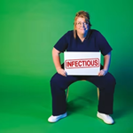 Nurse Georgie Carroll sits on a chair against a green background. She is wearing scrubs and holds a large sign that say 'Infectious'.