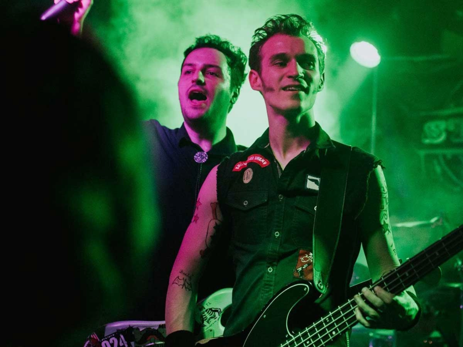 Two musicians perform on stage under green lighting. One is playing a black electric bass guitar and wearing a sleeveless shirt with patches, while the other stands slightly behind holding a microphone. The background features stage equipment and a smoky atmosphere, creating a vibrant live concert setting.