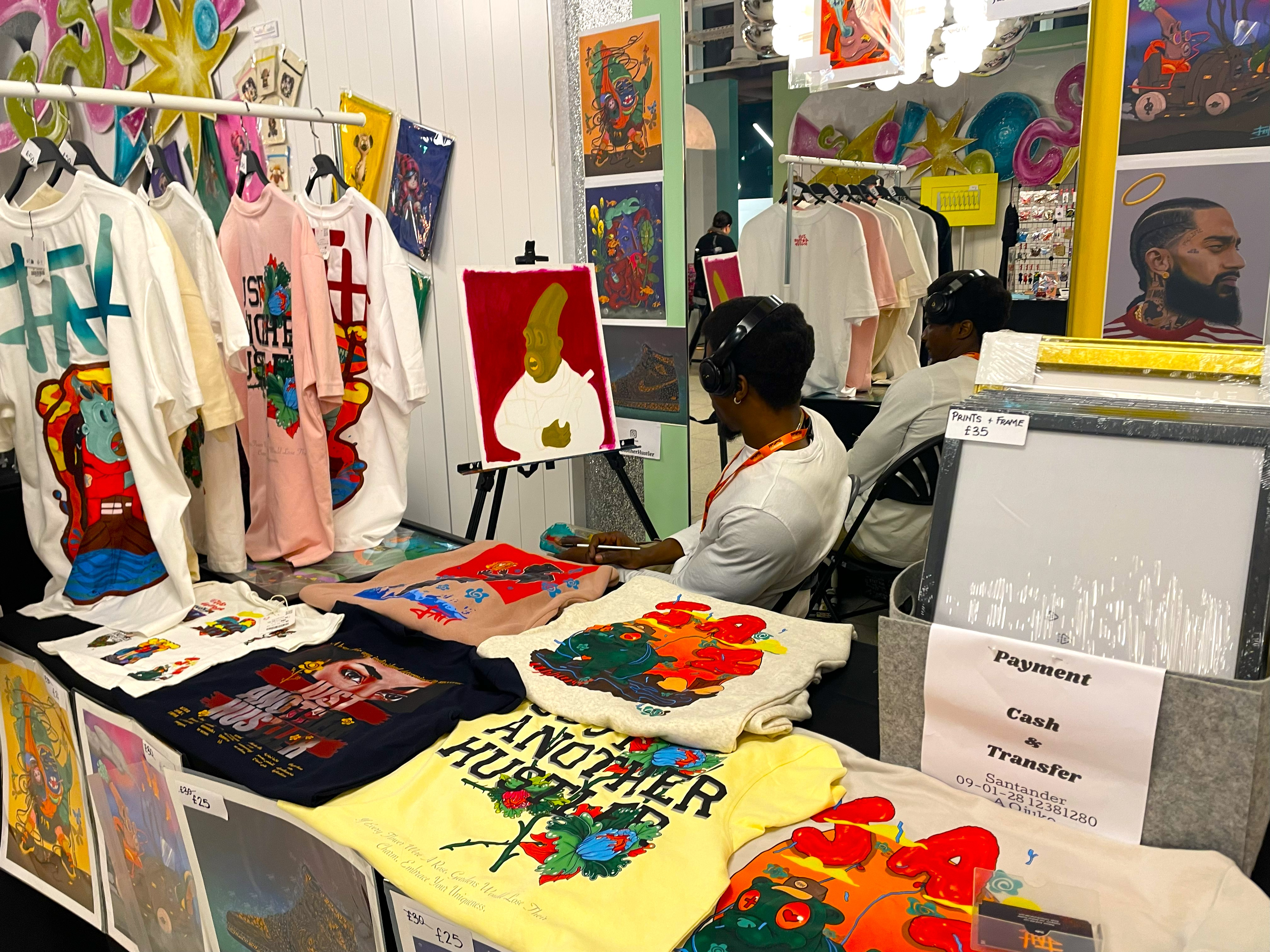 Pop-up market stall featuring vibrant printed T-shirts, framed artwork, and a painting on an easel. The table displays multiple colorful designs, and additional art pieces are hung on the wall behind.