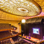 The Oval Hall at Sheffield City Hall. Grand auditorium with tiered seating and a large stage featuring a projection screen. The ornate ceiling has an intricate geometric design with a central stained-glass dome and a hanging light fixture. The stage is framed by black curtains, and the seating area includes multiple balconies with curved railings.