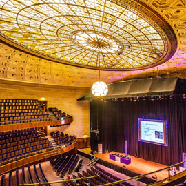 The Oval Hall at Sheffield City Hall. Grand auditorium with tiered seating and a large stage featuring a projection screen. The ornate ceiling has an intricate geometric design with a central stained-glass dome and a hanging light fixture. The stage is framed by black curtains, and the seating area includes multiple balconies with curved railings.