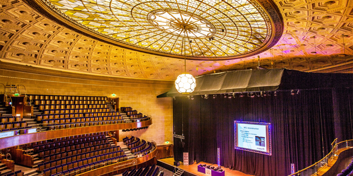 The Oval Hall at Sheffield City Hall. Grand auditorium with tiered seating and a large stage featuring a projection screen. The ornate ceiling has an intricate geometric design with a central stained-glass dome and a hanging light fixture. The stage is framed by black curtains, and the seating area includes multiple balconies with curved railings.