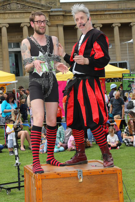 Two street performers stand on a wooden box during an outdoor event. One wears a black outfit with red and black striped socks and is wrapped in chains, holding a green prop. The other is dressed in a striking red and black costume with matching striped socks and boots. Behind them, a crowd watches near colourful tents and a large classical building, creating a lively festival atmosphere.