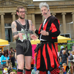Two street performers stand on a wooden box during an outdoor event. One wears a black outfit with red and black striped socks and is wrapped in chains, holding a green prop. The other is dressed in a striking red and black costume with matching striped socks and boots. Behind them, a crowd watches near colourful tents and a large classical building, creating a lively festival atmosphere.