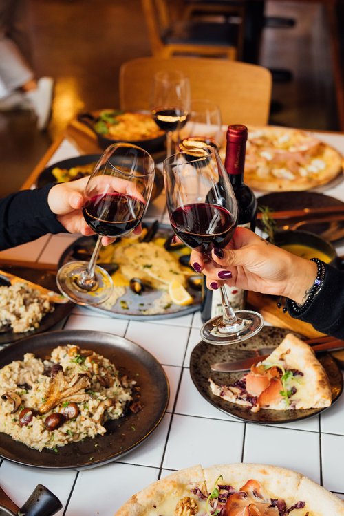 A table in a restaurant is laid out with a sumptuous meal. Two hands, from each side of the table, are holding glasses of red wine. 