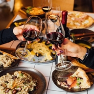 A table in a restaurant is laid out with a sumptuous meal. Two hands, from each side of the table, are holding glasses of red wine.