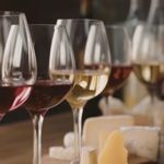 A row of wine glasses filled with different types of wine- ranging from deep red to pale white -stands on a wooden surface in soft, warm lighting. In front of and beside the glasses are assorted pieces of cheese in various shapes and textures, arranged casually as part of a tasting setup.