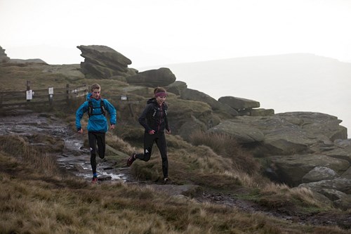 Two trail runners in outdoor gear run along a rocky path on a windswept hillside. The terrain is uneven with patches of grass and puddles, and large rock formations rise on the right side. A wooden fence and misty sky in the background suggest a remote, rugged landscape, possibly in a moor or upland area.