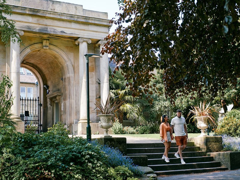 Couple walk down steps with the grand archway at the entrance to Sheffield Botanical Gardens in the background