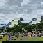 A busy outdoor fair on a large grassy field with groups of people sitting on hay bales and walking around. In the background are fairground rides, including two Ferris wheels and a tall yellow ride, along with colourful stalls and a striped tent. Trees line the edge of the field under a cloudy sky, creating a lively yet overcast atmosphere.
