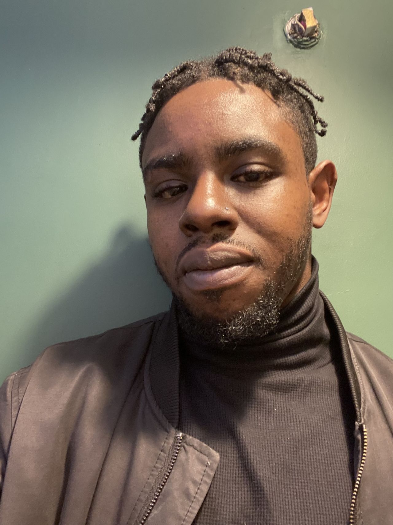 Person wearing a dark turtleneck sweater and a black jacket, standing against a plain green wall. A small round fixture is visible on the wall near the top right of the image.