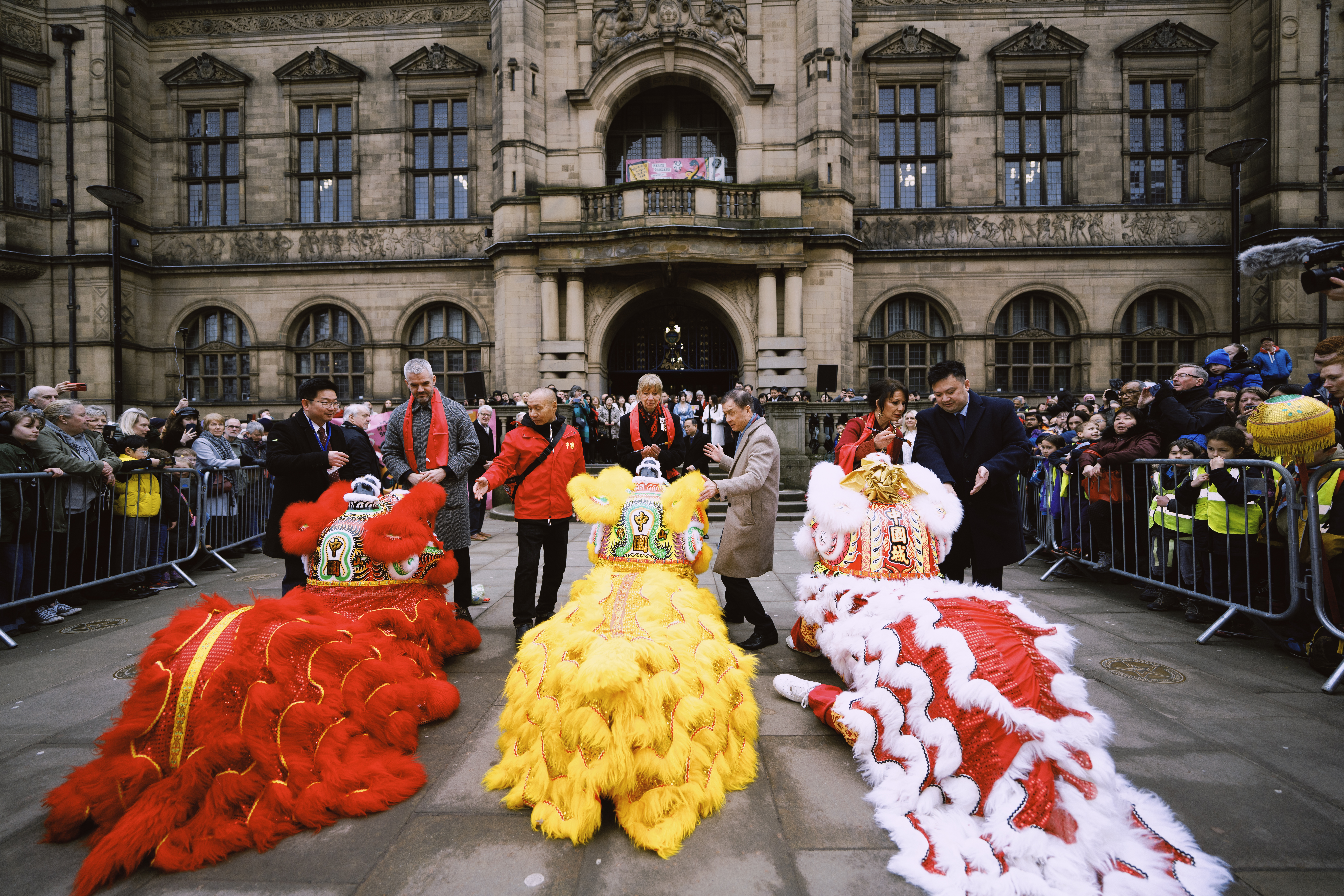 Three lion dance costumes—red, yellow, and white—kneeling on the ground in front of a historic building entrance. Several people in festive attire stand nearby, and a crowd watches from behind barriers.