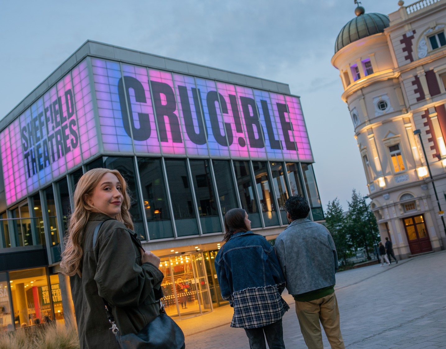 Sheffield Theatres’ Crucible building glowing pink at dusk, showing people arriving for an evening performance in the city centre.