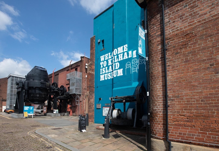 The entrance to the Kelham Island Museum. On the right is a blue-painted wall with 'Welcome To Kelham Island Museum' on it. In the background you can see a huge Bessemer Converter that was used in the production of steel.