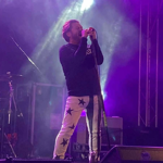 A performer stands on stage singing into a microphone, wearing a black long-sleeve top and white pants decorated with large black stars. Bright purple and white stage lights create a dramatic backdrop, with drums and equipment visible in the background.