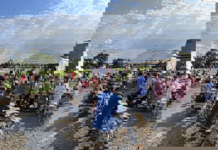 A sunny outdoor scene at a beer festival with groups of people sitting at wooden picnic benches on a paved area beside a large grassy field. A white marquee tent is set up in the background with people gathered around it. The sky is bright with scattered clouds.