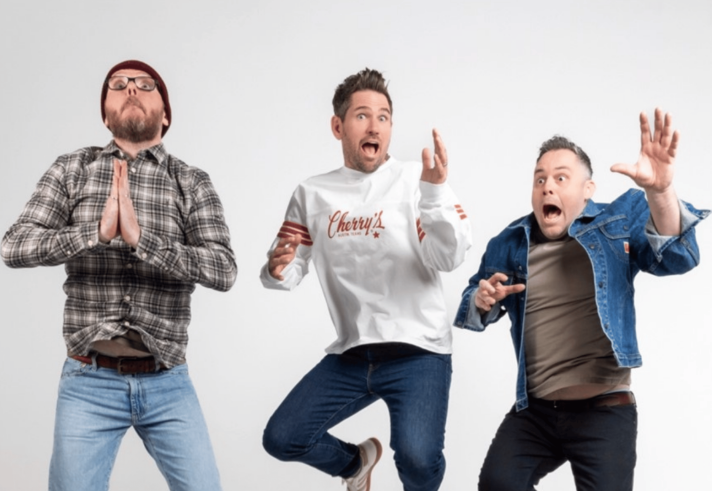 Three people are mid-jump against a plain white background. The person on the left wears a plaid shirt and jeans with hands pressed together in a prayer-like gesture. The person in the center wears a white sweatshirt with “Cherry’s” written on it and dark jeans, with arms bent and fingers pointing upward. The person on the right wears a denim jacket over a gray shirt and dark pants, with one arm raised and the other bent forward in an expressive pose.