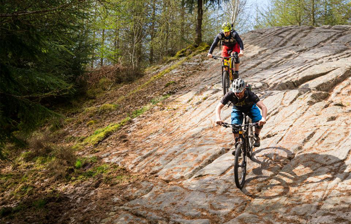 Two people on mountains bikes ride down a rocky slope in the countryside.