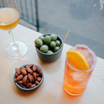A bowl of olives, a bowl of nuts, a glass of wine and a cocktail on a table.