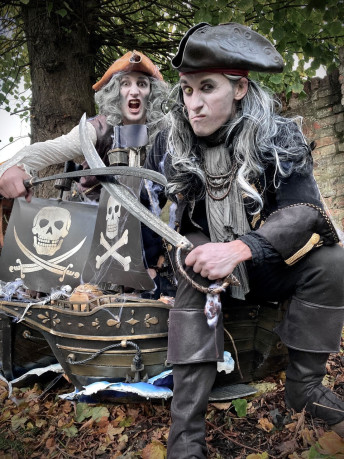 Two men dressed as pirates pose next to a model of a pirate ship.