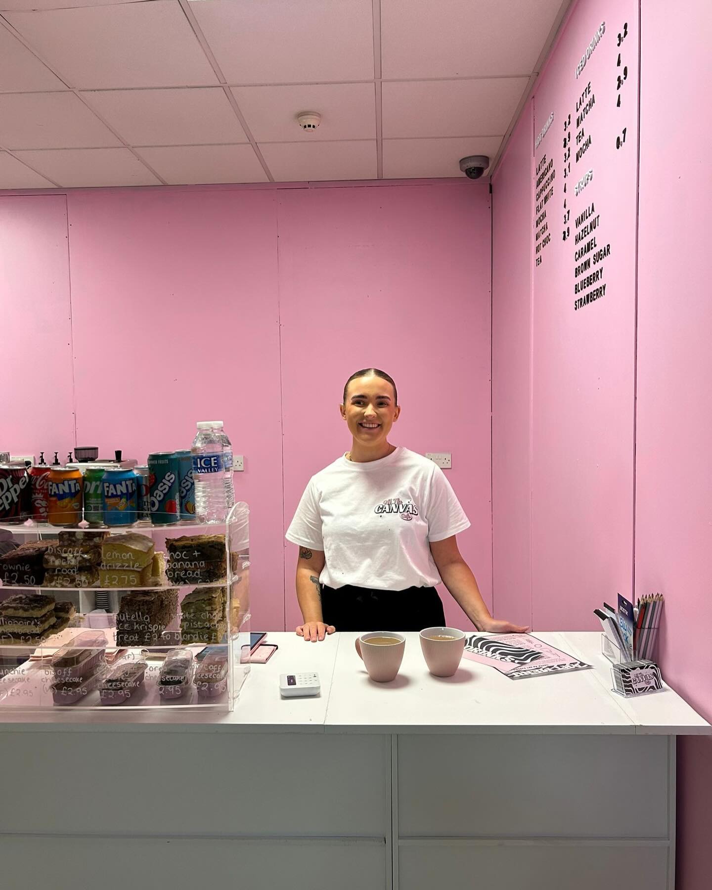A person standing behind a white counter in a pastel pink room, with shelves of drinks and snacks to the side and a menu displayed on the wall.