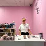 A person standing behind a white counter in a pastel pink room, with shelves of drinks and snacks to the side and a menu displayed on the wall.