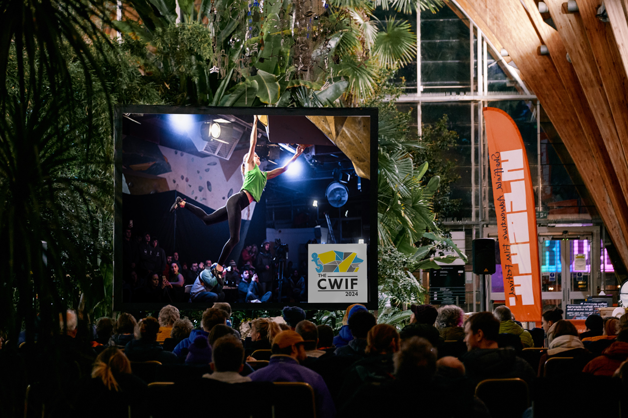 A large indoor screen displays a climber in a green top hanging from a bouldering wall during the CWIF 2024 event. The screen is set up in a spacious venue filled with lush green plants and wooden beams, with rows of seated spectators watching. An orange banner with text stands to the right, and bright lighting highlights the modern glass structure in the background.